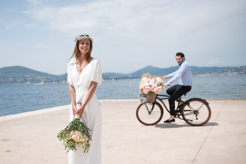Scène de mariage captée sur le vif : la mariée marche au bord de l’eau, bouquet à la main, pendant que le marié la rejoint à vélo, instant spontané et lumineux en reportage photo de mariage.