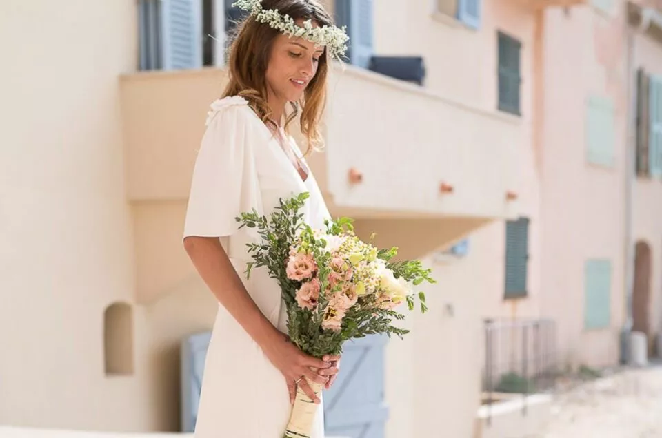 Mariée à Saint-Tropez tenant son bouquet, photographiée dans une rue aux façades pastel du Sud, lumière douce et naturelle.