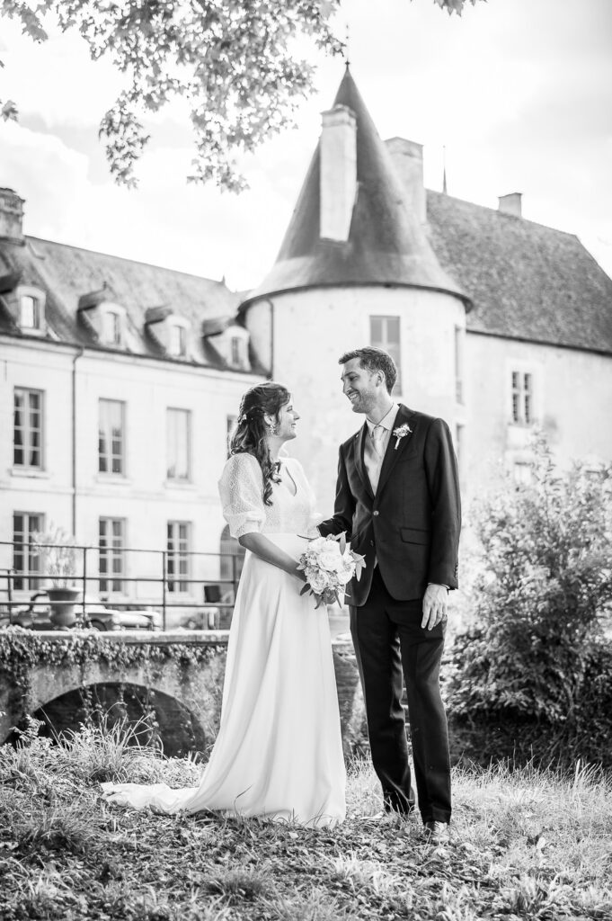 Couple de mariés se tenant la main devant le château de Riceys-Bas, portrait en noir et blanc dans le parc du château, moment complice et naturel en reportage de mariage.