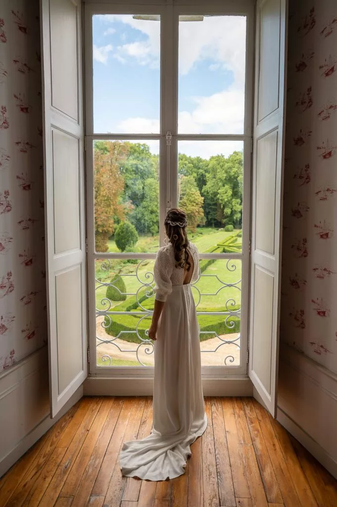 Mariée de dos devant une grande fenêtre du château de Riceys-Bas, robe élégante et vue sur le parc du château, lumière douce et ambiance intime de mariage.