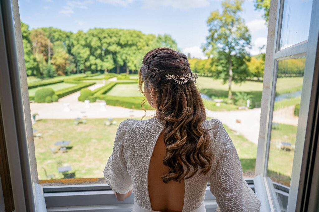 Mariée regardant le parc du château de Riceys-Bas depuis une fenêtre, coiffure élégante et robe dos nu, lumière naturelle et instant de calme avant le mariage.