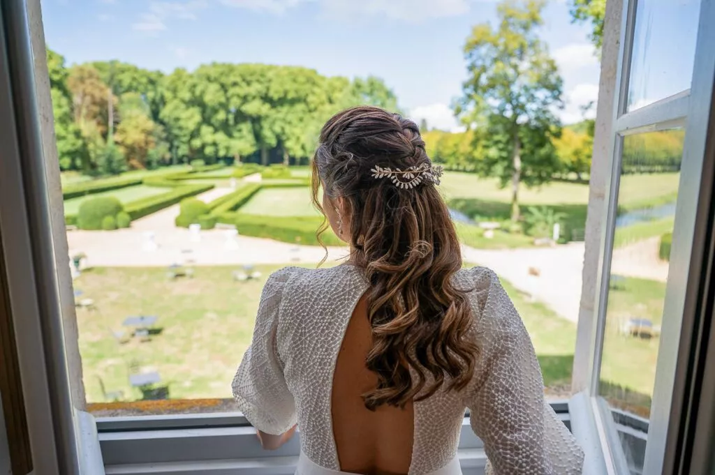 Mariée regardant le parc du château de Riceys-Bas depuis une fenêtre, coiffure élégante et robe dos nu, lumière naturelle et instant de calme avant le mariage.