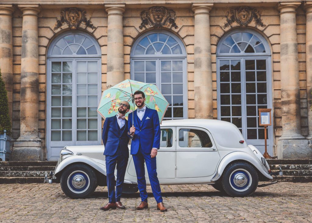 Couple de mariés en costumes bleus sous un parapluie coloré devant une voiture ancienne, reportage photo de mariage élégant devant un château.