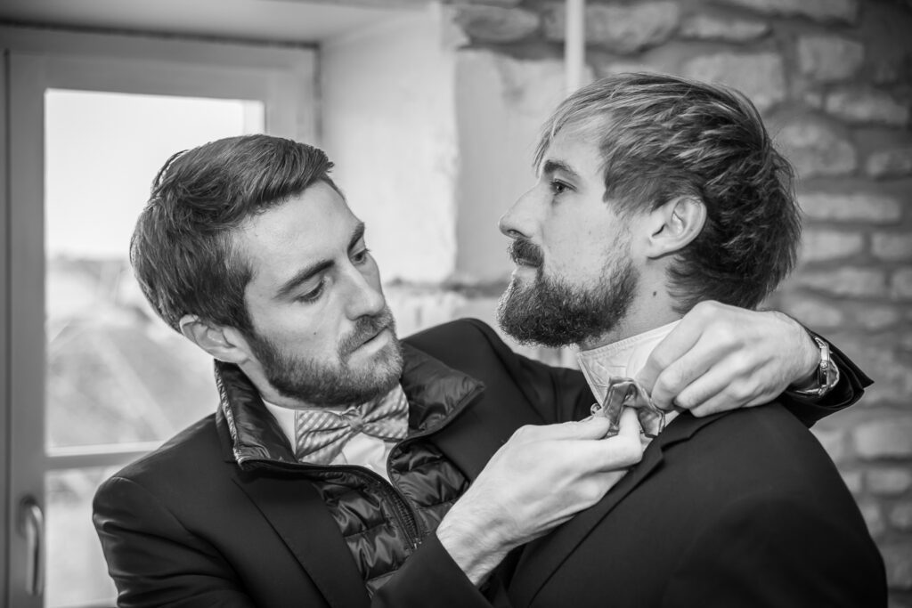 Préparatifs du marié, un proche ajuste le nœud papillon dans une atmosphère intime, scène captée sur le vif en noir et blanc.
