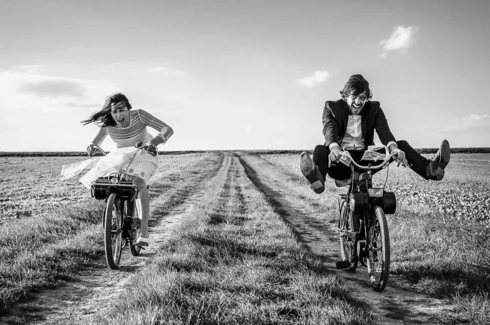 Les mariés roulent chacun sur un Solex sur un chemin de campagne, dans un moment joyeux et spontané, en noir et blanc.