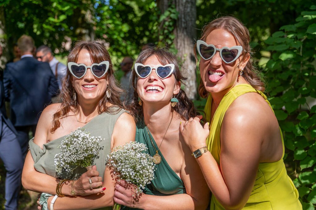 Trois invitées souriantes portent des lunettes en forme de cœur lors d’un mariage en extérieur.