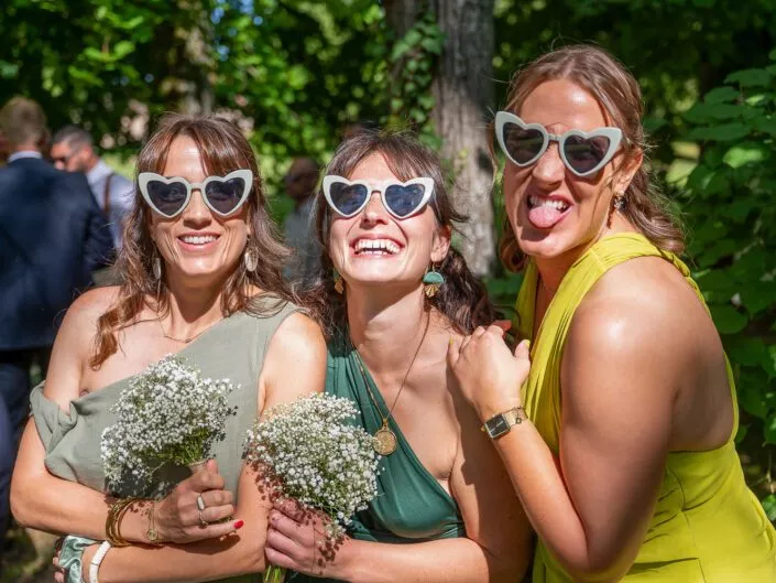 Trois invitées souriantes portent des lunettes en forme de cœur lors d’un mariage en extérieur.