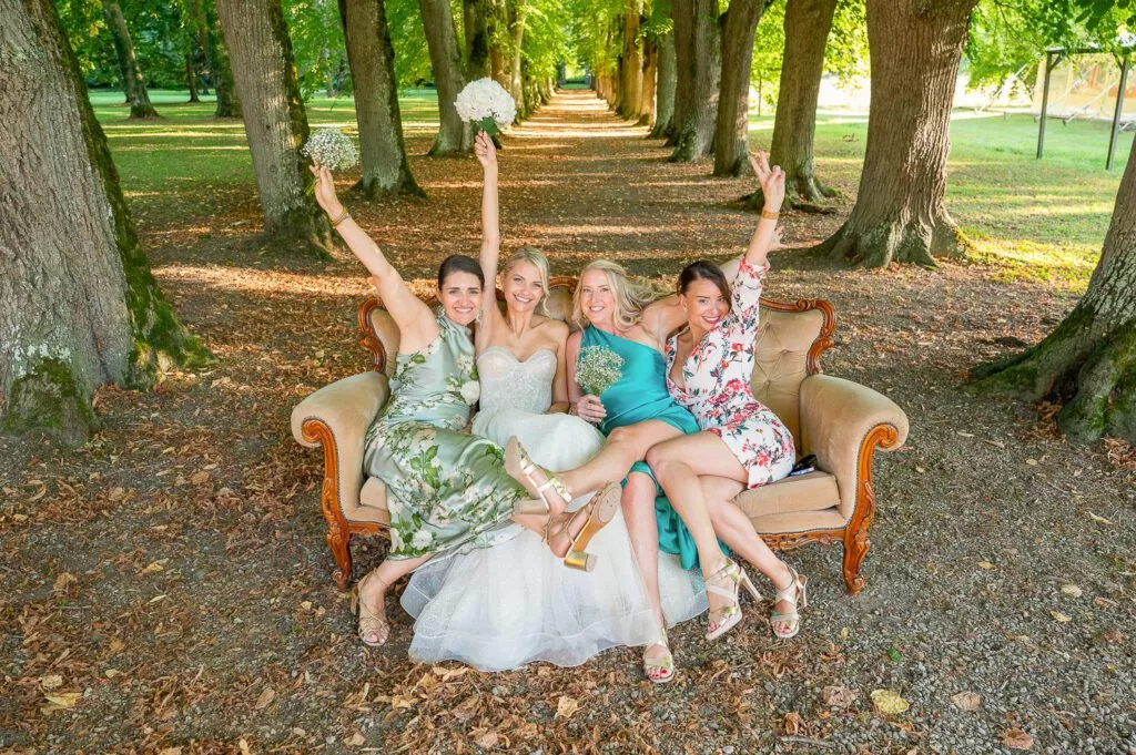 La mariée et ses amies célèbrent ensemble sur un canapé, devant une allée d’arbres menant au domaine du mariage.