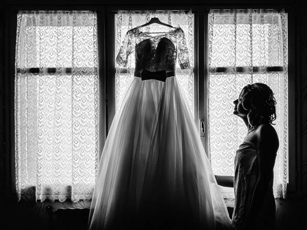 La mariée observe sa robe suspendue devant la fenêtre, moment de calme et d’émotion avant la cérémonie, photographie en noir et blanc.