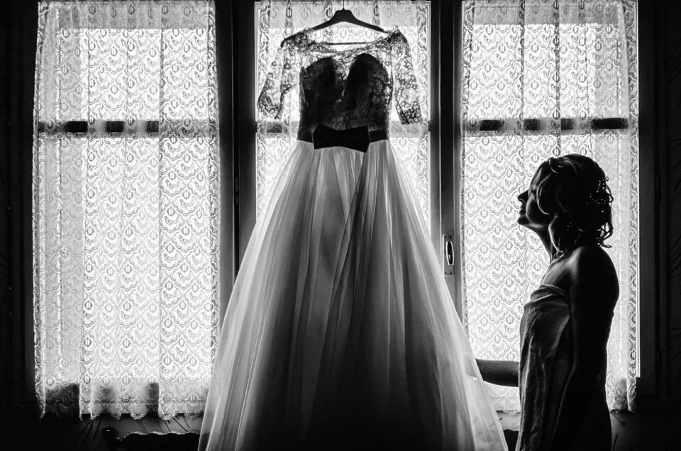 La mariée observe sa robe suspendue devant la fenêtre, moment de calme et d’émotion avant la cérémonie, photographie en noir et blanc.