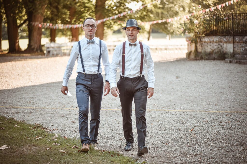 Deux invités en tenue élégante avec bretelles et nœud papillon marchant dans le parc du Domaine de Vauluisant près de Troyes lors d’un reportage photo de mariage.